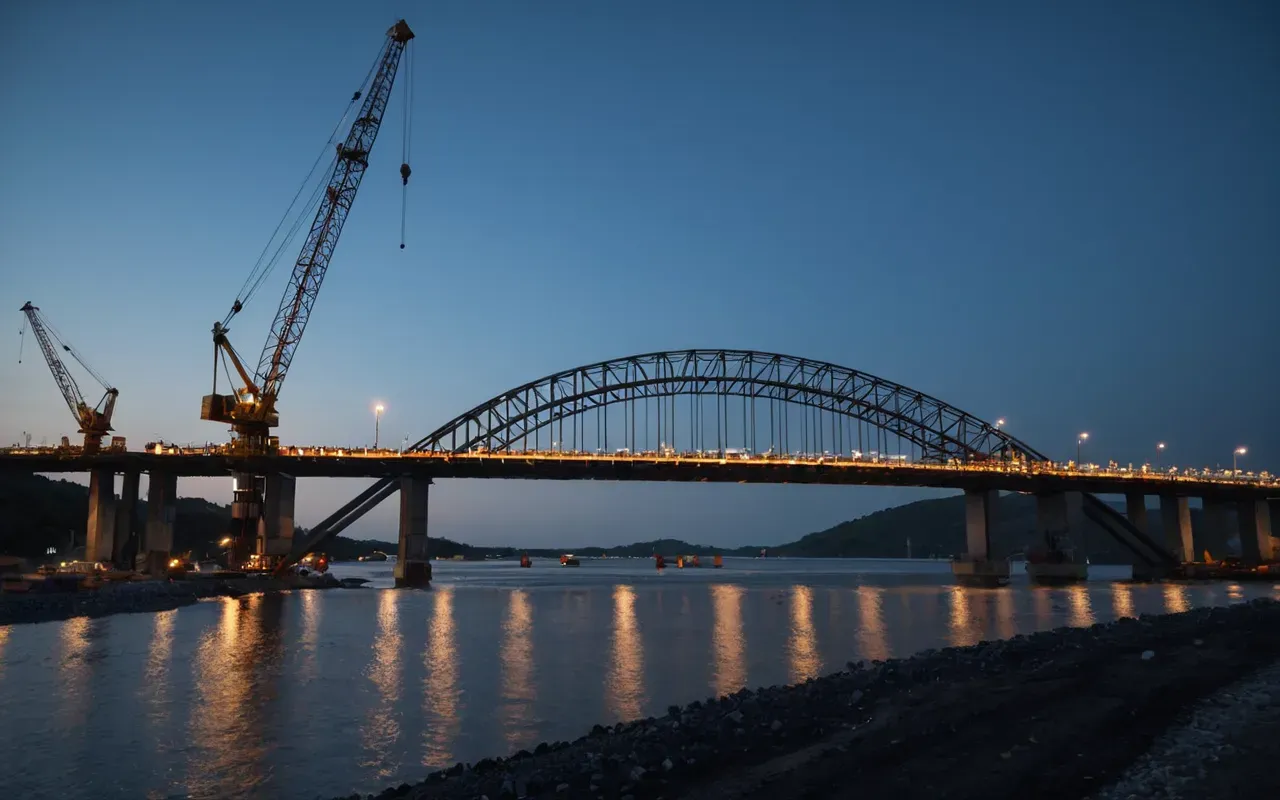 A wide shot of a massive infrastructure bridge under construction at dusk, with large cranes and concrete pouring equipment visible against a dark blue sky
